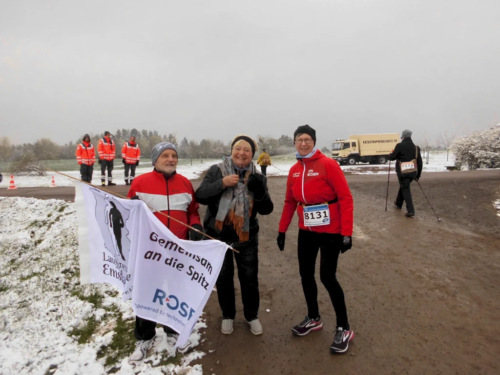 Läufer:innen vor Schnee-Landschaft bei einem Winterrennen, tragen Startnummern und Banner, im Hintergrund ein LKW.