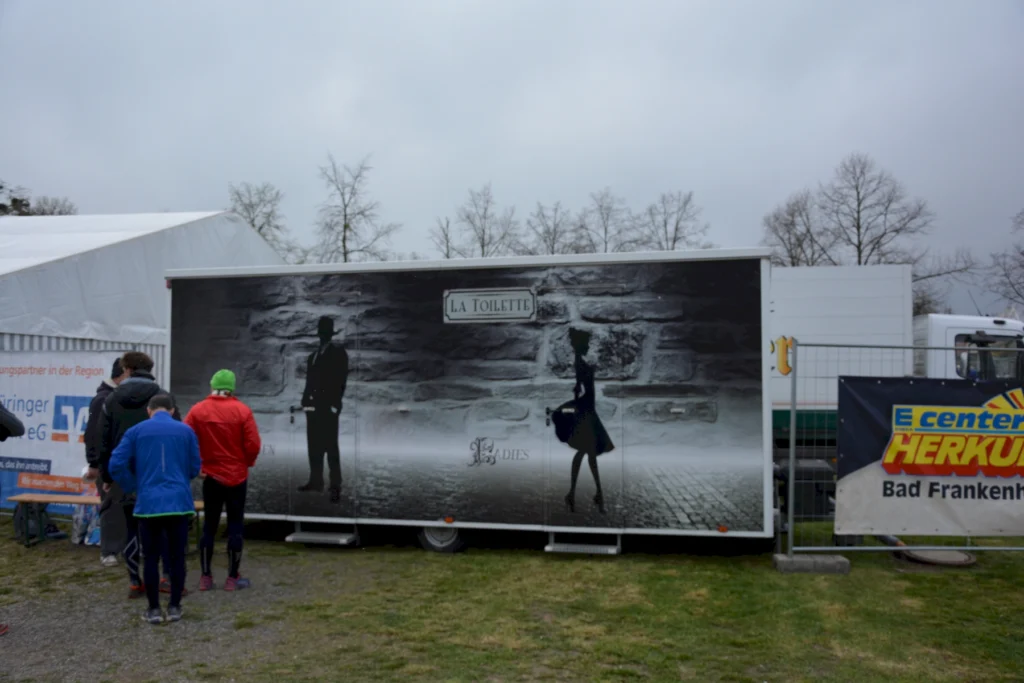 Toilettenwagen mit schwarz-weißer Wanddekoration, Figuren eines Mannes und einer Frau, umringt von Menschen im Freien.