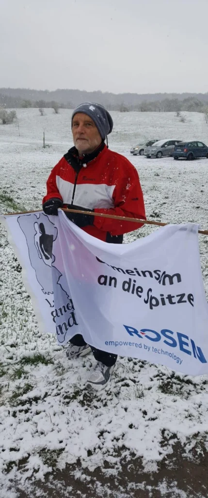 Ein Mann im Schnee hält eine Fahne mit dem Text Gemeinsam an die Spitze. Im Hintergrund parken Autos.