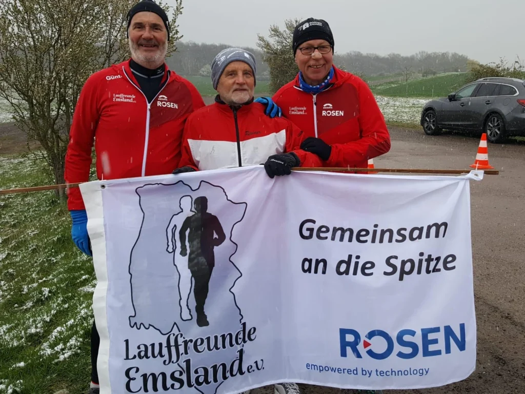 Drei Personen in roten Jacken halten ein Banner des Vereins Lauffreunde Emsland. Schneebedeckte Landschaft im Hintergrund.