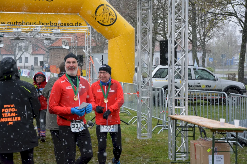 Läufer im Schnee beim Zieleinlauf eines Rennens, tragen rote Jacken und Medaillen, Hintergrund Autohaus-Barbarossa-Banner.