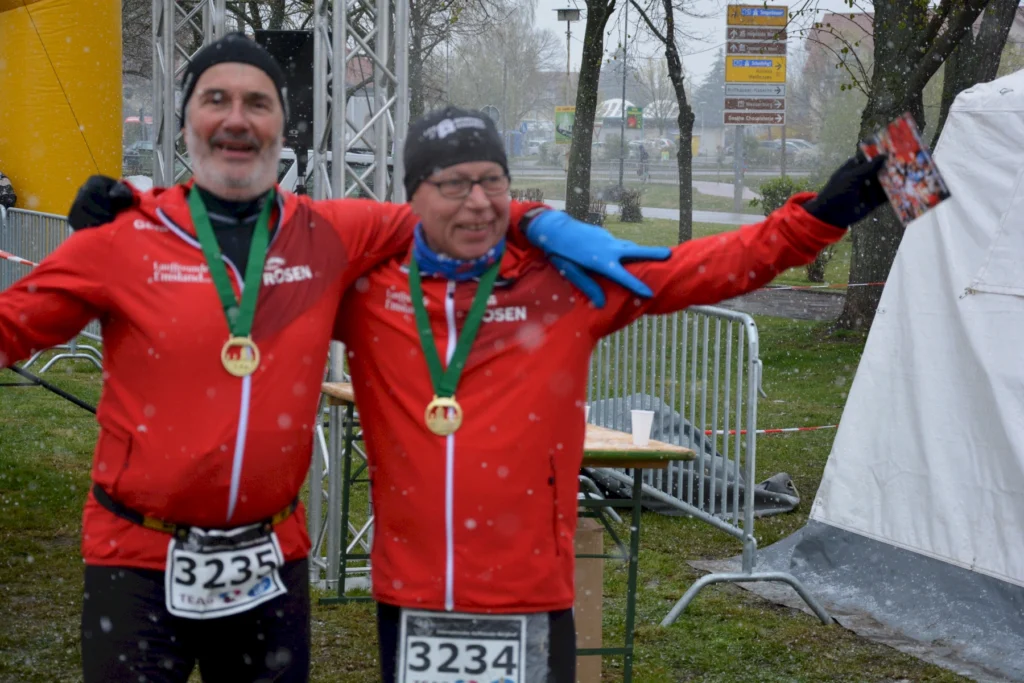 Zwei Läufer im roten Trikot jubeln mit Medaillen bei einem Winterlauf im Freien mit leichtem Schneefall.