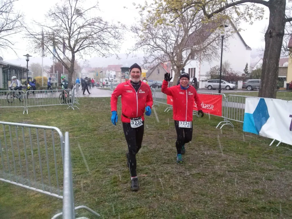 Zwei Läufer in roten Jacken beim Lauf durch einen verschneiten Park nahe einer Wohnsiedlung, Zielbereich in Sicht.