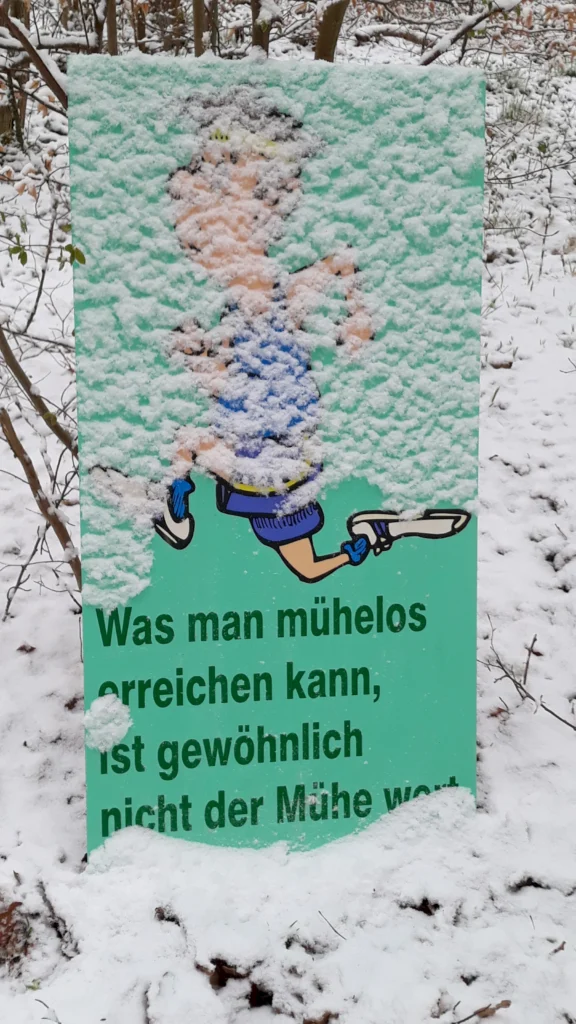 Schneebedecktes Schild im Wald mit Spruch: Was man mühelos erreichen kann, ist gewöhnlich nicht der Mühe wert.
