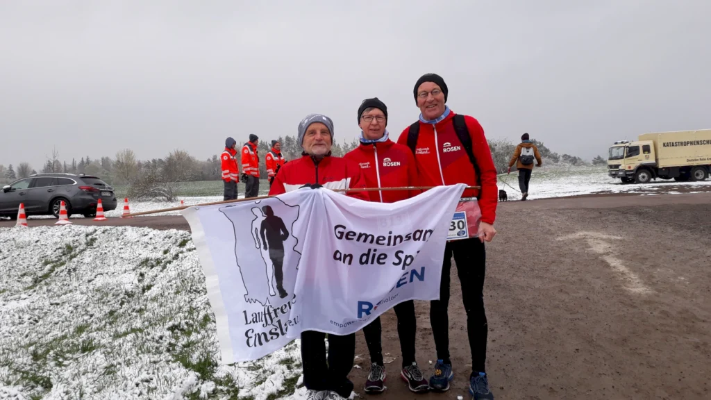 Drei Läufer mit Banner im Winter bei einem Laufereignis, umgeben von schneebedeckter Landschaft und Rotkreuz-Helfern.