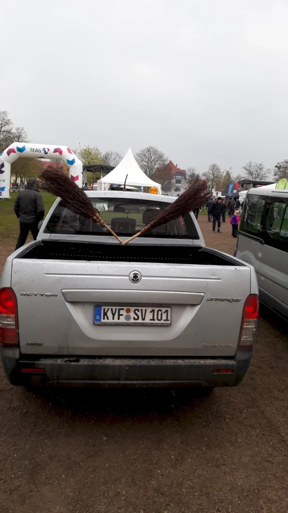 Silberner Pick-up mit zwei Reisigbesen auf der Ladefläche bei einem Outdoor-Event. Zelte und Menschen im Hintergrund.