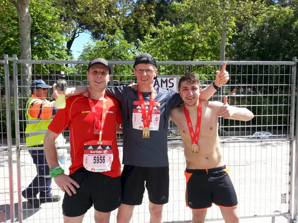 Drei Läufer nach dem Wettkampf, stolz mit Medaillen um den Hals und Getränken in der Hand, stehen vor einem Zaun. Im Hintergrund ist ein Sicherheitsmitarbeiter zu sehen. Die Szene vermittelt Freude und Erfolg nach einem Marathon.