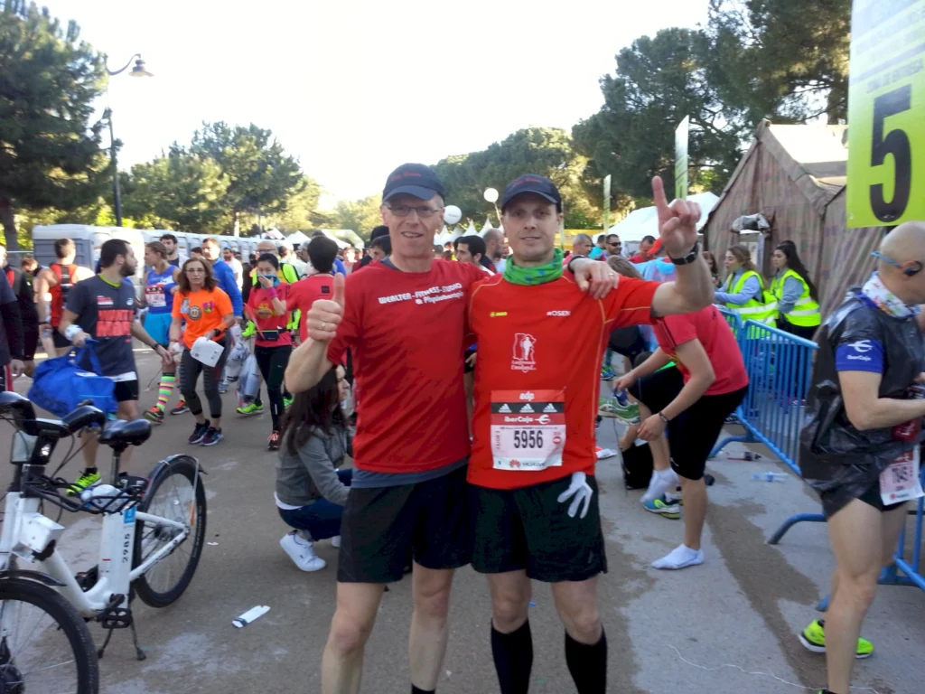 Zwei Teilnehmer eines Marathons posieren lächelnd in roten Shirts vor der Startlinie, während im Hintergrund zahlreiche Zuschauer und andere Läufer zu sehen sind. Die Atmosphäre ist festlich und sportlich, mit bunten Outfits und einem Fahrrad in der Nähe.