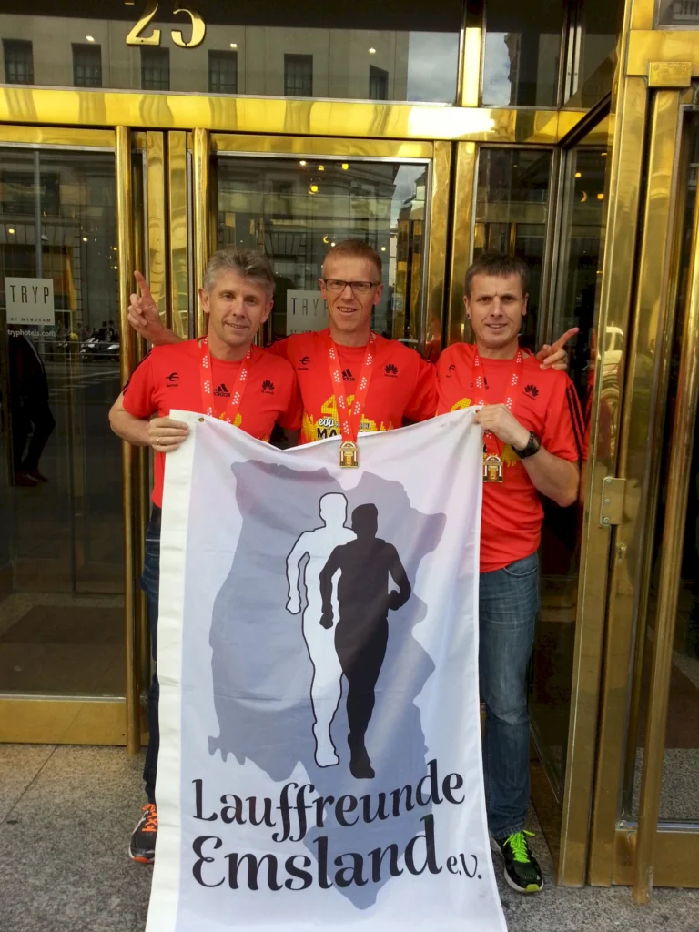 Drei Läufer in roten T-Shirts mit Medaillen stehen vor einem goldenen Eingang und halten ein Banner der Lauffreunde Emsland e.V. in der Hand.