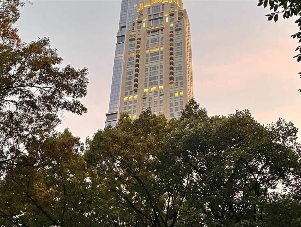 Hochhaus in der Stadt umgeben von Bäumen bei Sonnenuntergang, mit erleuchteten Fenstern und einem klaren Himmel im Hintergrund.