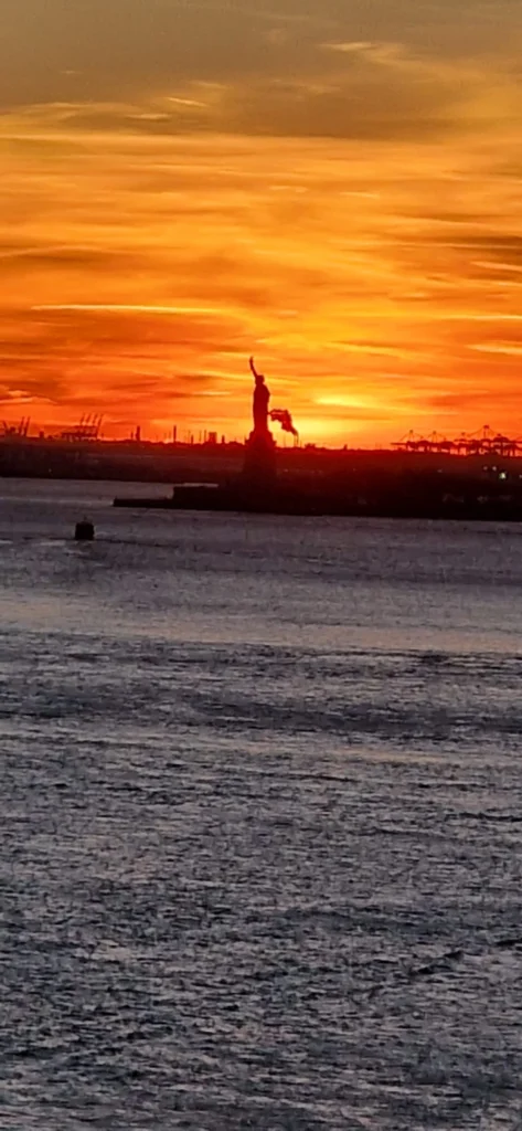 Die Statue der Freiheit vor einem dramatischen Sonnenuntergang über dem Wasser, mit orangefarbenem Himmel und Silhouetten von Hafenkränen im Hintergrund.