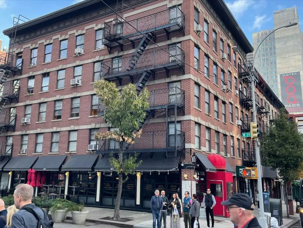 Ziegelstein-Gebäude mit Feuerwehrleitern in einer belebten Stadtstraße, umgeben von Fußgängern und Geschäften, im Hintergrund ein Hotel-Schild.