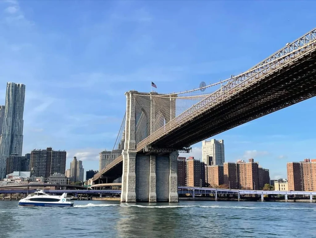 Brooklyn Bridge in New York City mit Blick auf die Skyline, ein Boot fährt auf dem East River unter der Brücke hindurch.