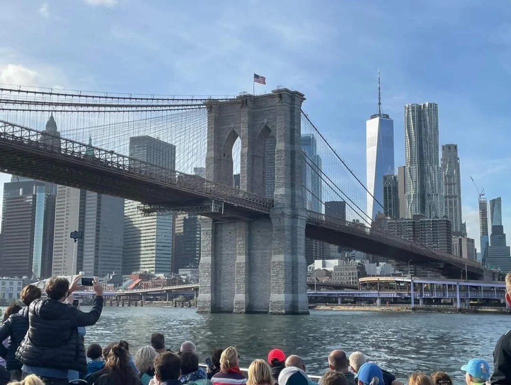 Blick auf die Brooklyn Bridge mit der Skyline von Manhattan im Hintergrund, während Touristen auf einem Boot die Sehenswürdigkeit fotografieren. Die beeindruckende Architektur der Brücke und die modernen Wolkenkratzer, darunter das One World Trade Center, sind gut sichtbar.