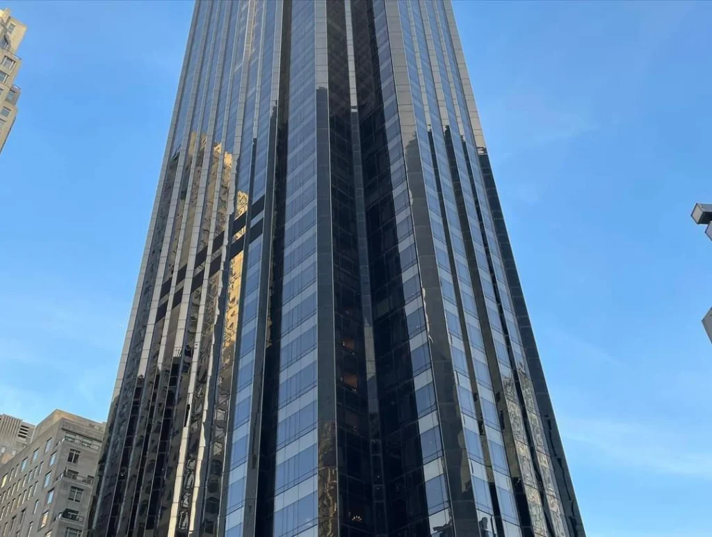 Hochhaus mit spiegelnder Glasfassade vor blauem Himmel, umgeben von urbaner Architektur.