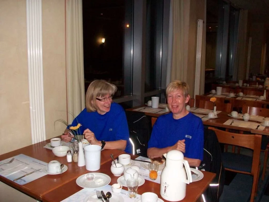 Zwei Personen in blauen T-Shirts frühstücken lächelnd an einem Tisch in einem Café mit leerem Geschirr im Hintergrund.