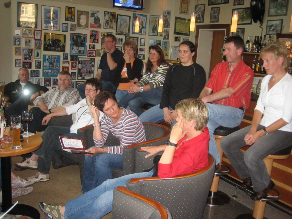 Personengruppe bei informellem Treffen in einer Bar mit Wänden voller Fotos, entspannt bei Getränken sitzend.