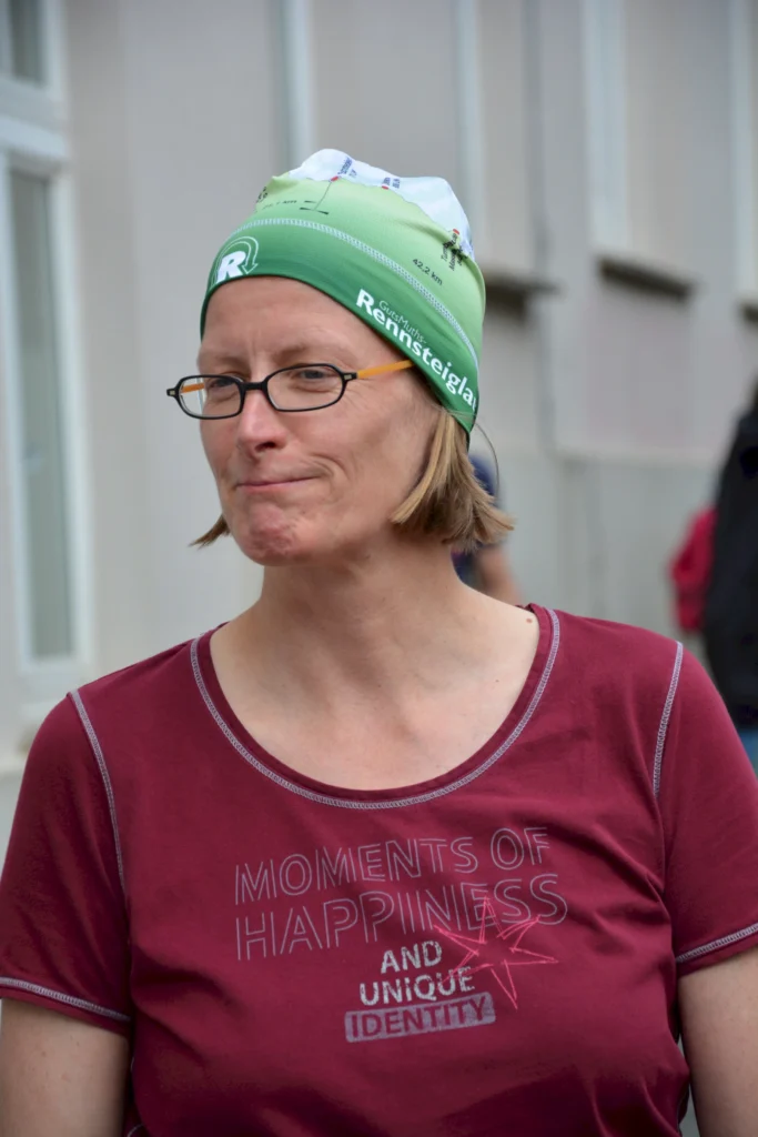 Person mit Brille und Rennsteiglauf-Mütze in rotem Shirt mit Aufdruck „Moments of Happiness“ auf der Straße.