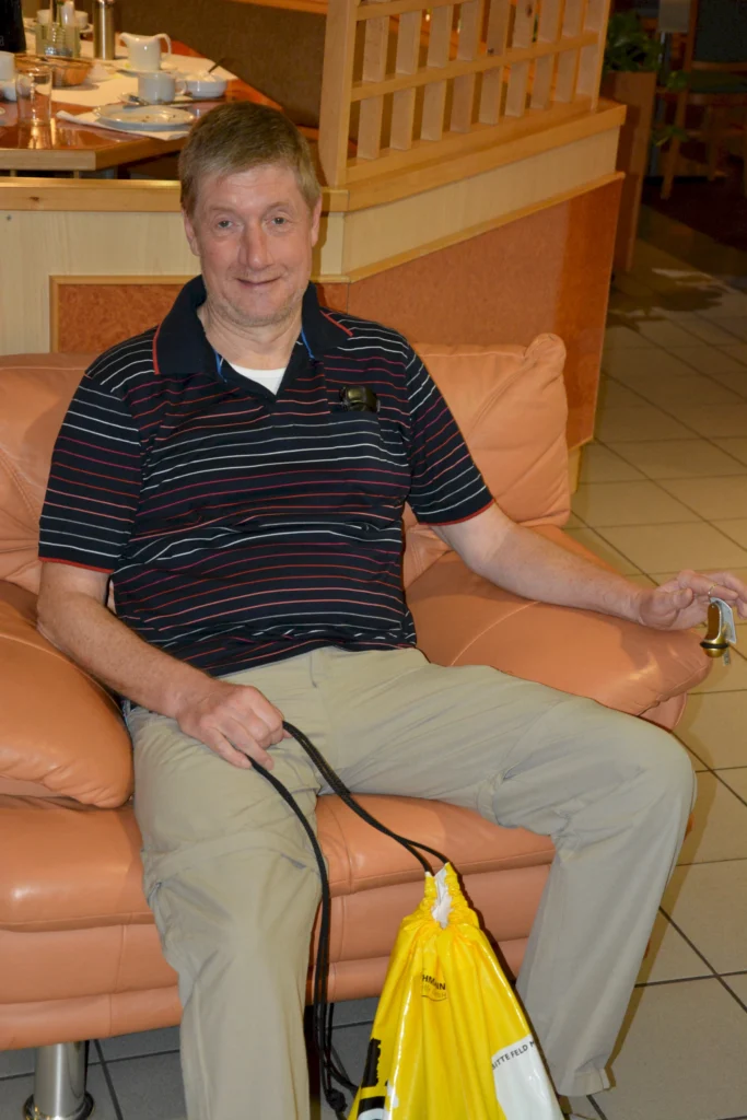 Mann in Streifenpolo sitzt auf einem Sofa mit gelbem Rucksack und Schlüsseln in der Hand, entspannt in einem Café.