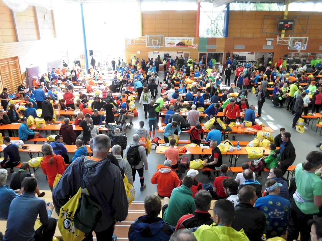 Große Gruppe von Menschen in bunter Sportkleidung bei Veranstaltung in Sporthalle, sitzend an Tischen.
