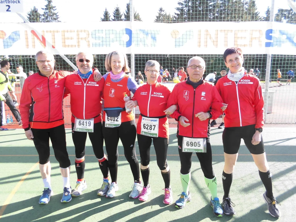 Läufergruppe in roten Sportjacken posiert vor einem INTERSPORT-Banner bei sonnigem Wetter auf einem Sportplatz.