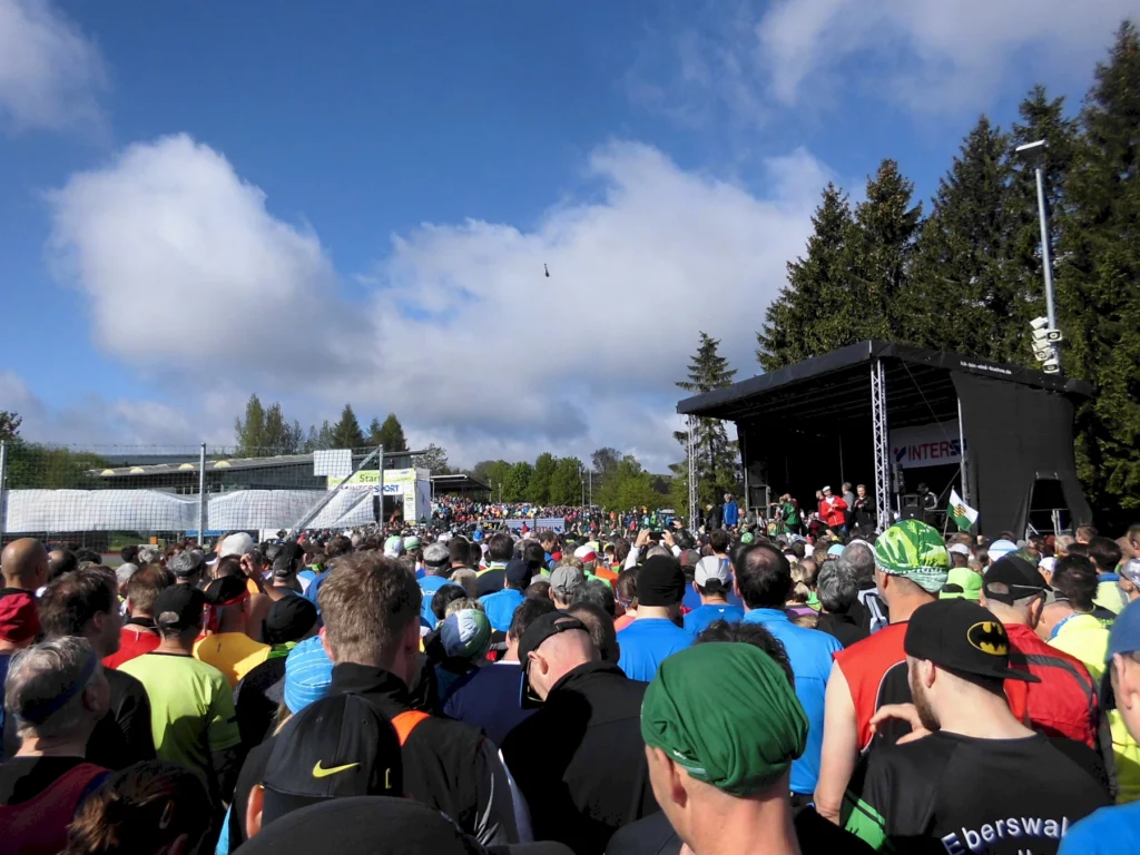 Läufer versammeln sich am Start eines Marathons bei sonnigem Wetter, während Redner auf einer Bühne sprechen.