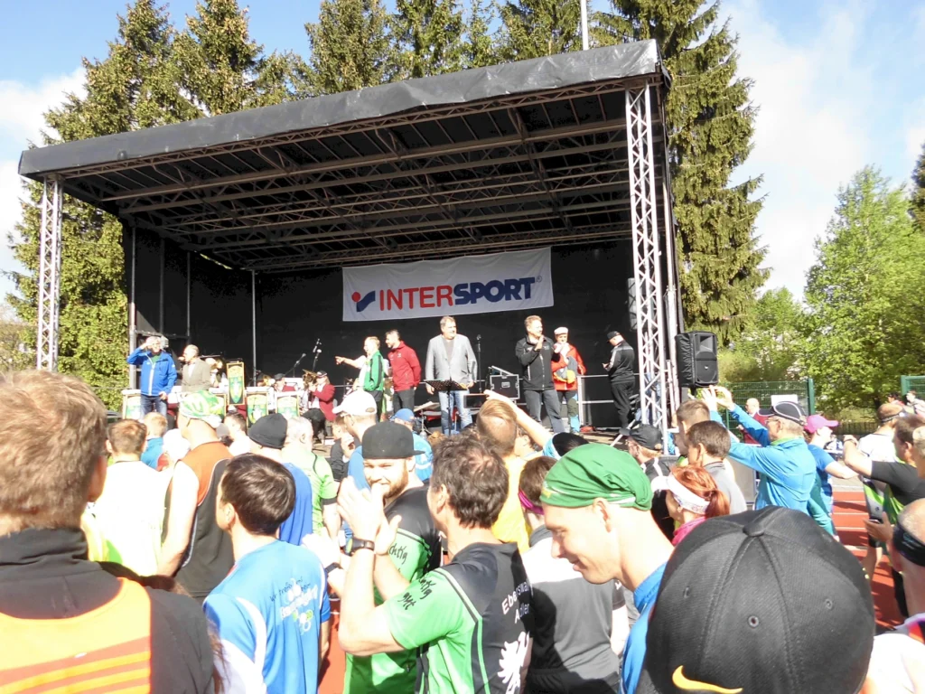 Läufer versammeln sich vor der Bühne bei einer Sportveranstaltung im Freien, Intersport-Banner im Hintergrund.