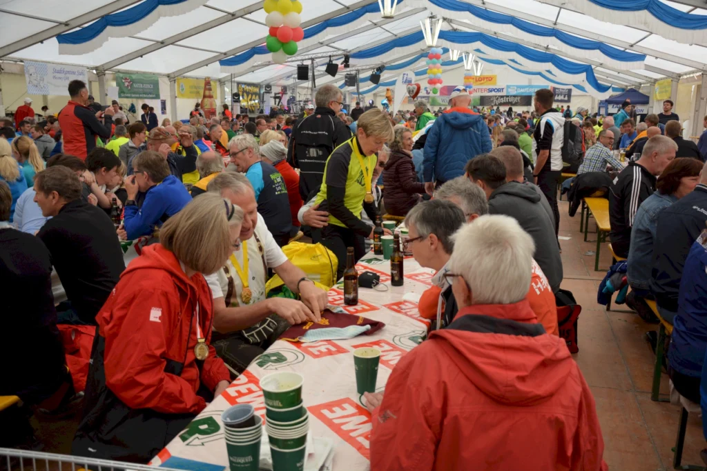Menschen feiern in einem Festzelt, sitzen an langen Tischen und unterhalten sich bei Getränken und Essen.