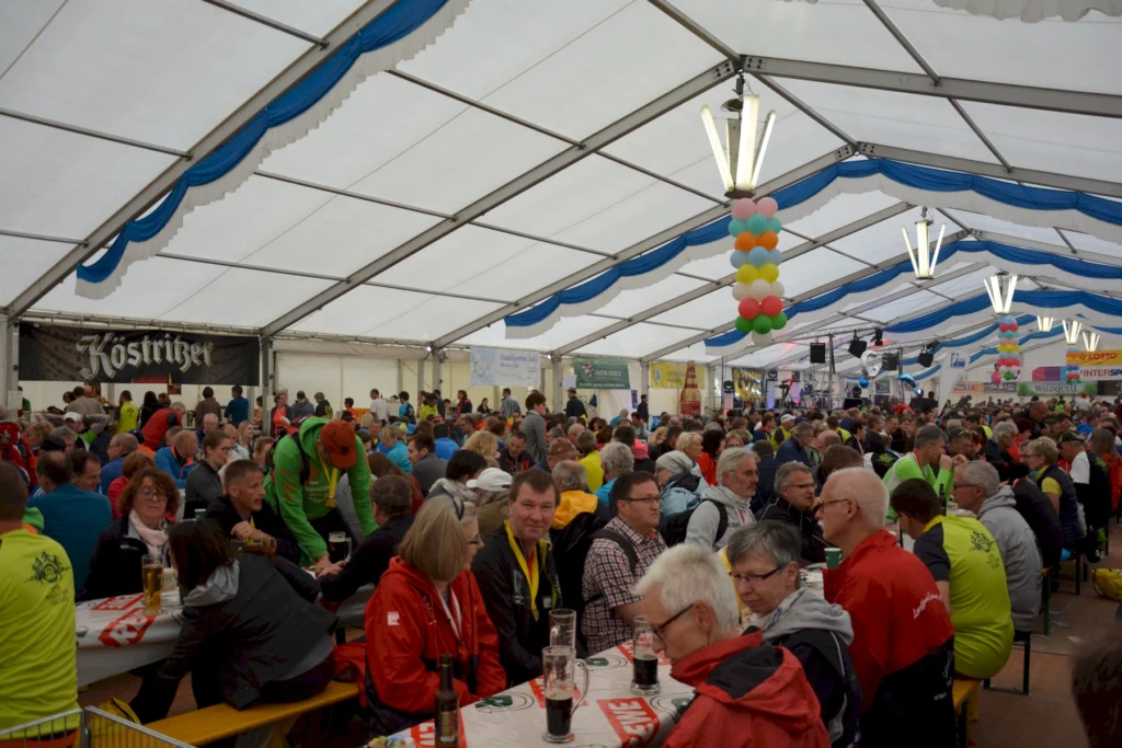 Großes Festzelt mit vielen Menschen, die an Tischen sitzen und Bier trinken, geschmückt mit bunten Luftballons.
