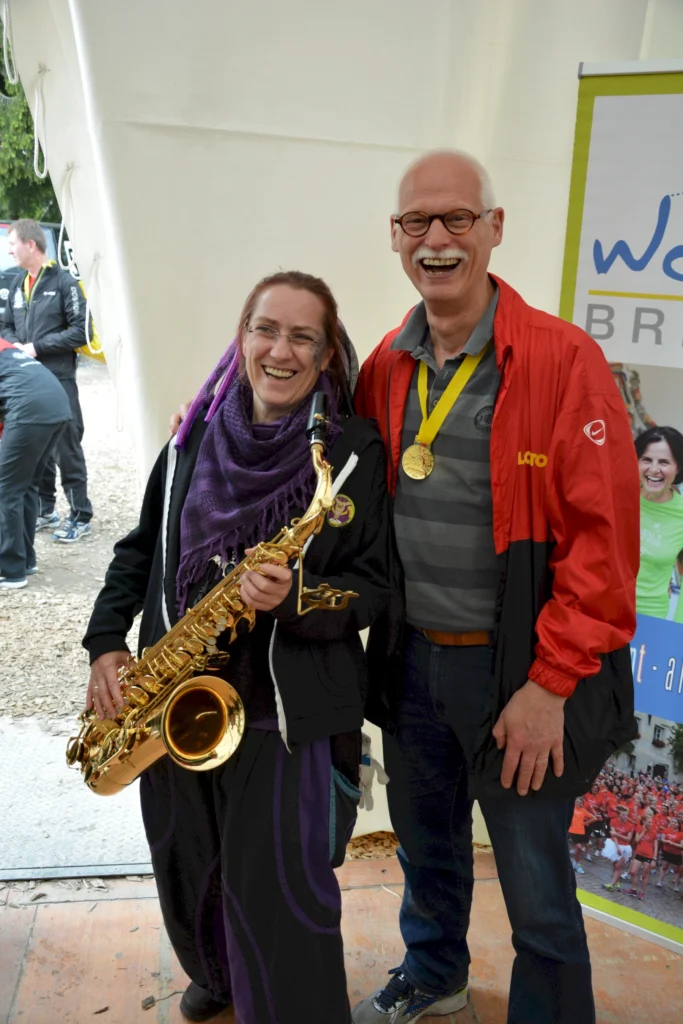 Zwei lachende Personen mit Saxophon und Medaille bei einem Event, vor einem Werbeplakat in einem Zelt.