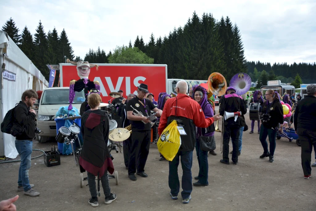 Blaskapelle spielt im Freien auf einem Festival, umgeben von Besuchern und einem großen roten Lastwagen.