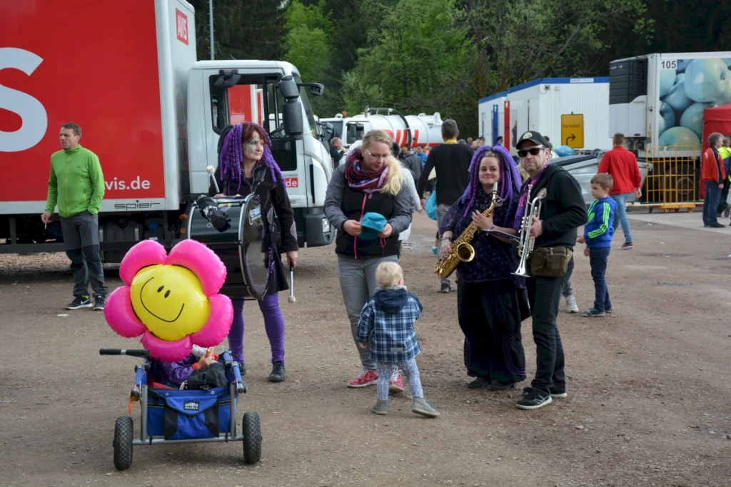 Menschen in bunten Kostümen mit Instrumenten auf einem Fest, Kind im Vordergrund mit rotem Blumenballon.