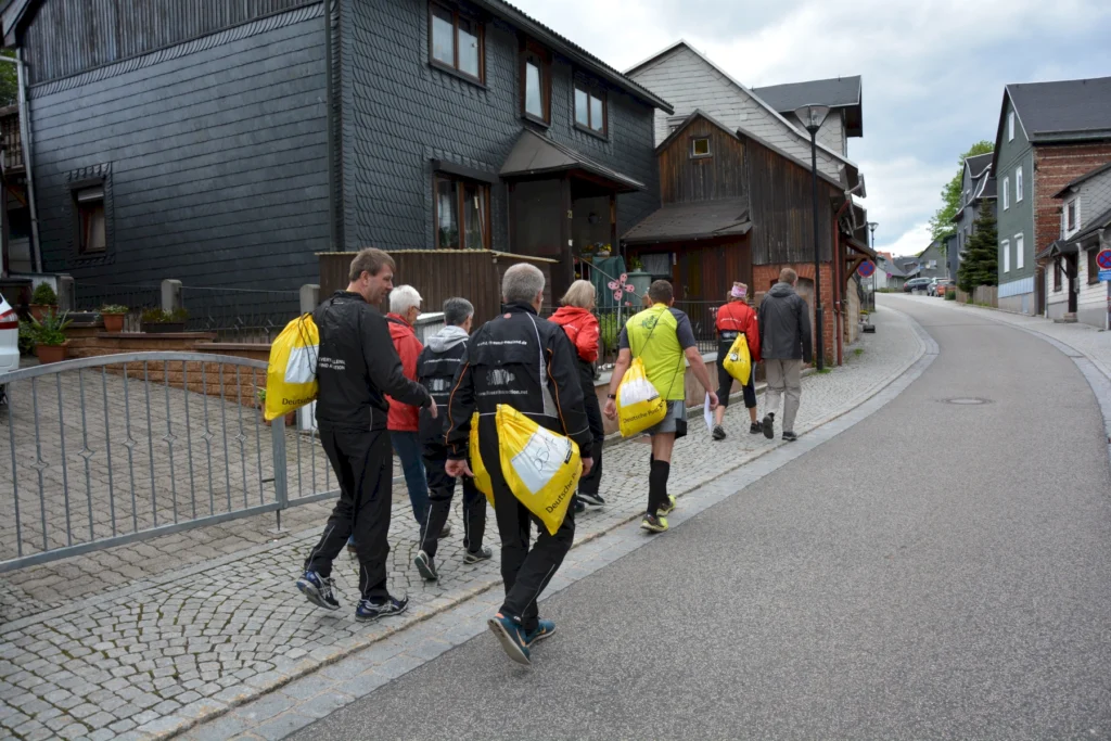 Gruppe von Menschen wandert auf gepflastertem Weg durch Dorf, trägt gelbe Taschen.