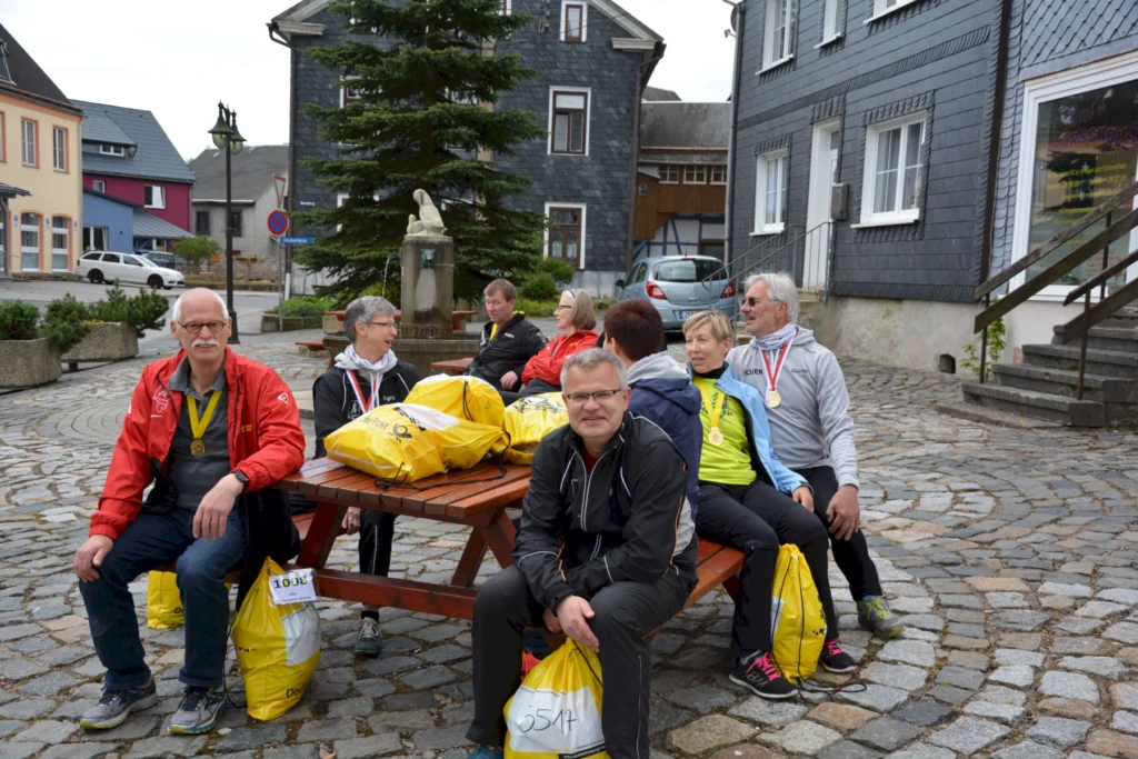 Gruppe älterer Menschen mit Medaillen sitzt auf Bänken in einem Dorfplatz, umgeben von alten Häusern.