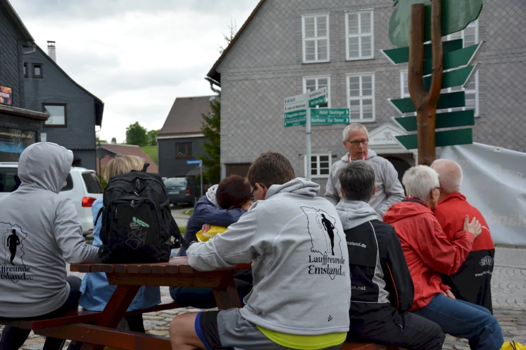 Gruppe von Menschen sitzt an einem Holztisch im Freien und hört einem Mann in einem grauen Hoodie zu. Gebäude im Hintergrund.