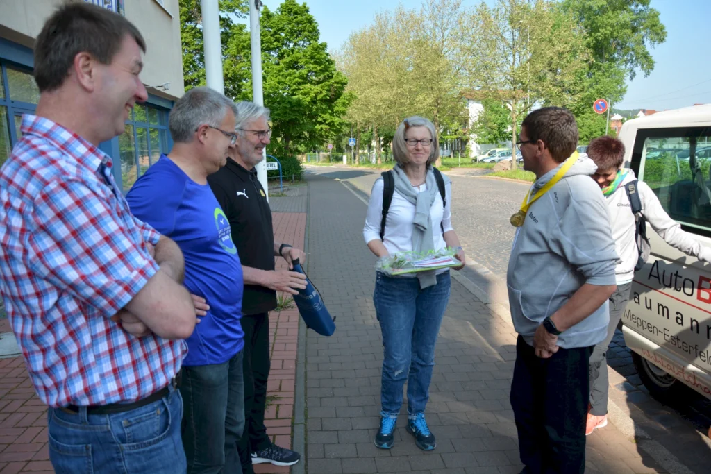 Gruppe von Menschen unterhält sich freundlich im Freien neben einem Auto, eine Frau hält ein Geschenk in der Hand.