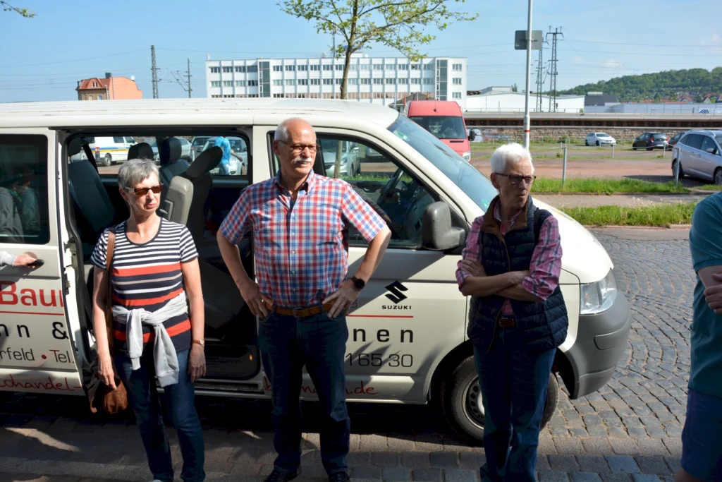 Personen stehen neben einem weißen Kleinbus auf einer Straße, im Hintergrund städtische Gebäude und grüne Landschaft.
