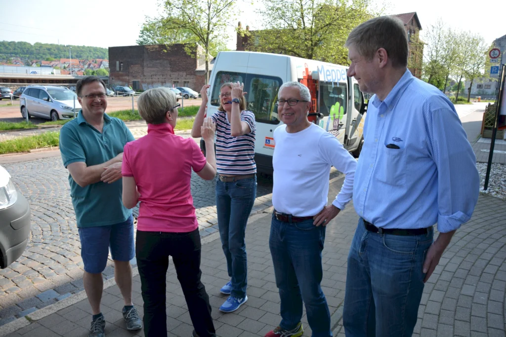 Gruppe von fünf Personen, die lachend im Freien steht, mit einem Kleinbus im Hintergrund bei sonnigem Wetter.