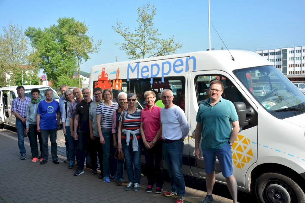 Gruppe von Menschen steht neben einem Kleinbus mit der Aufschrift Meppen bei sonnigem Wetter auf dem Gehweg.