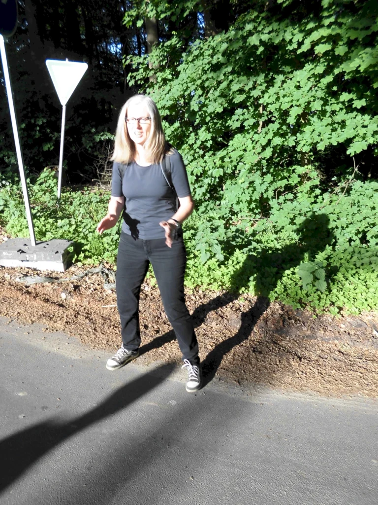 Frau in schwarzem Oberteil und schwarzen Hosen steht auf einem Weg, umgeben von grünen Bäumen und Sträuchern, mit einem Verkehrsschild im Hintergrund. Sie wirkt fröhlich und gestikuliert, während die Sonne auf sie scheint.