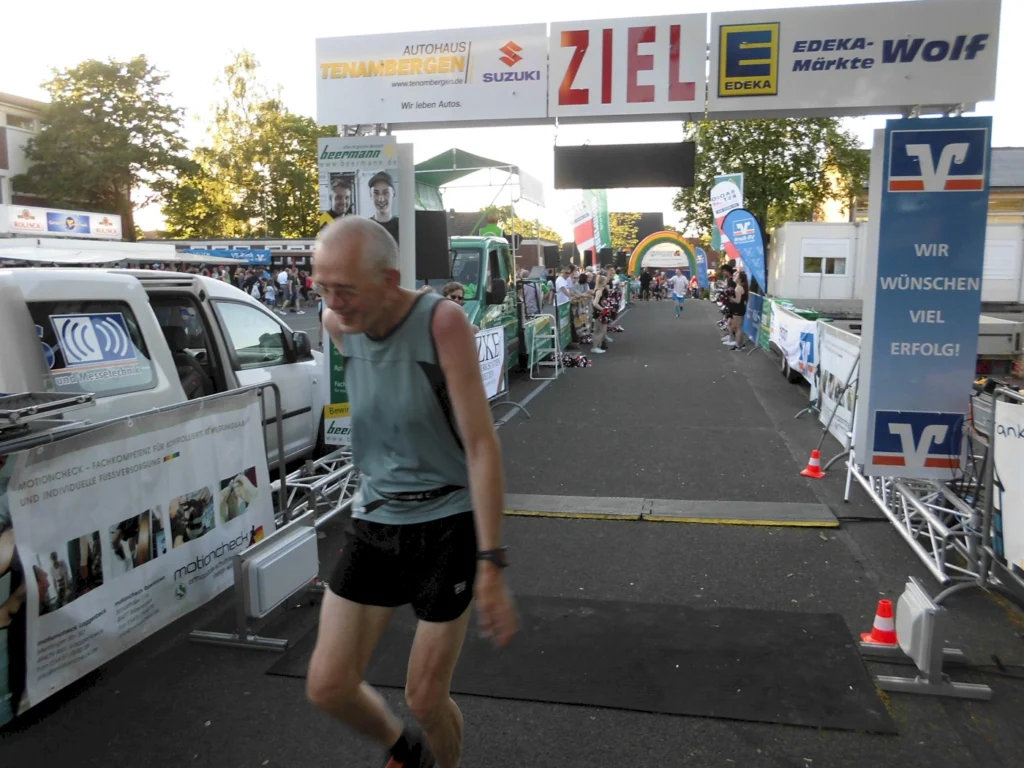 Ein älterer Läufer überquert die Ziellinie bei einem Sportevent, umgeben von Zuschauern und Werbebannern. Im Hintergrund sind verschiedene Sponsorenlogos sichtbar, darunter EDEKA und Suzuki.