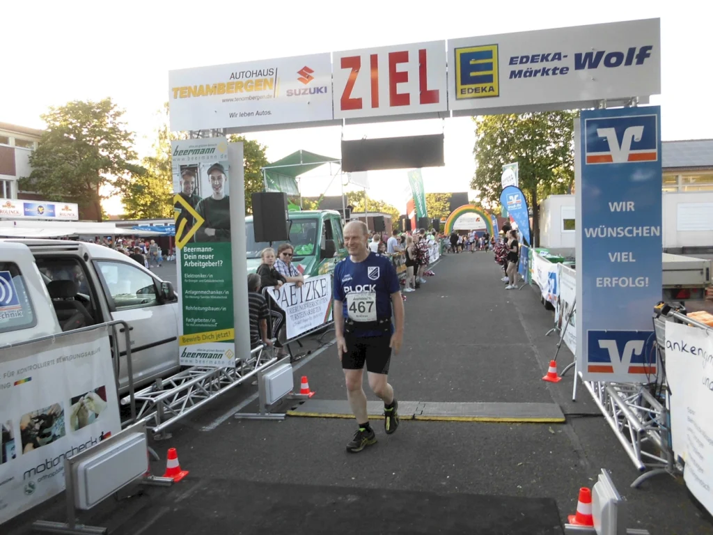 Läufer überquert die Ziellinie eines Wettlaufs, umgeben von Zuschauern und Sponsorenbanner. Im Hintergrund sind verschiedene Stände und ein Zielbogen sichtbar, was auf eine sportliche Veranstaltung hinweist.