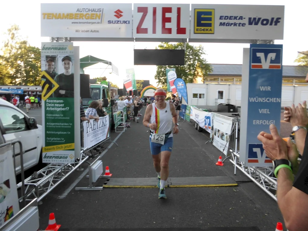 Läufer überquert die Ziellinie eines Wettkampfs, umgeben von jubelnden Zuschauern und Sponsorenbanner, bei einem Sportevent in Deutschland.