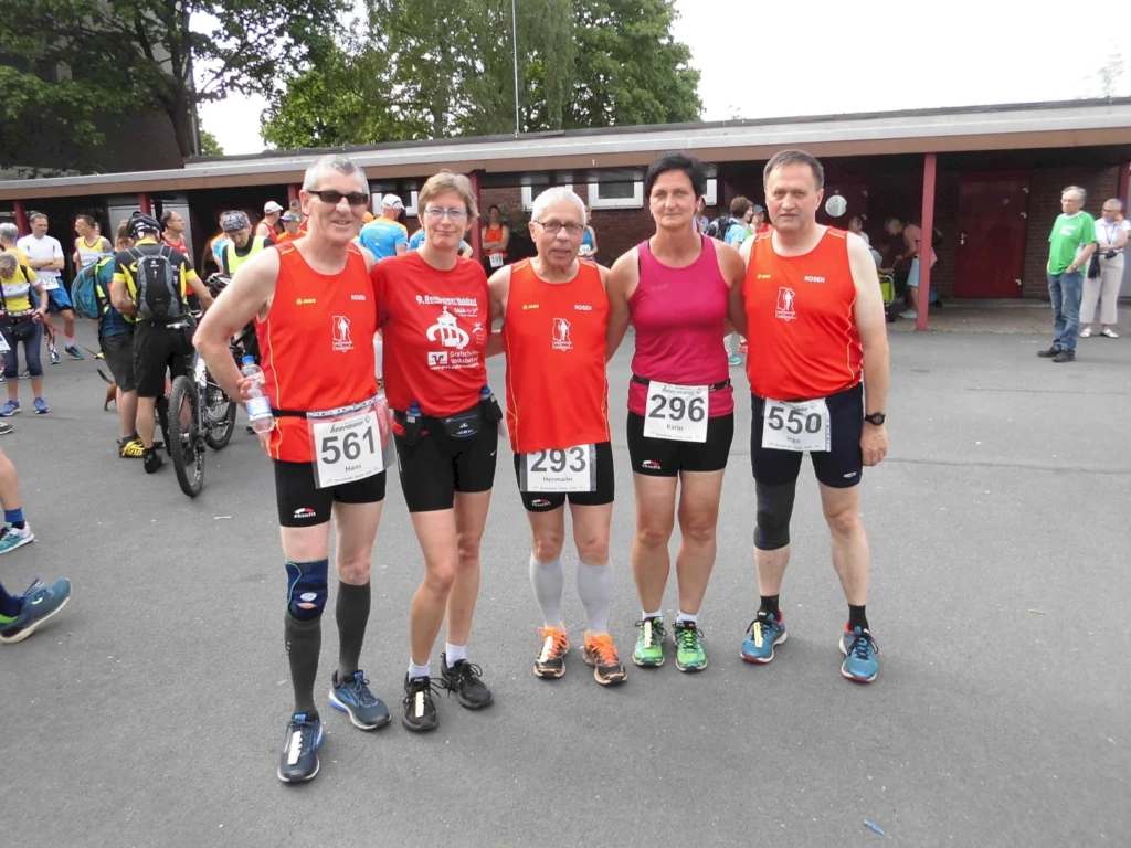 Fünf Sportler in Laufbekleidung stehen vor einer Gruppe von Zuschauern und anderen Teilnehmern. Die Athleten tragen rote Trikots mit Nummern und bereiten sich auf ein Rennen vor. Im Hintergrund sind Zuschauer und weitere Sportler zu sehen.