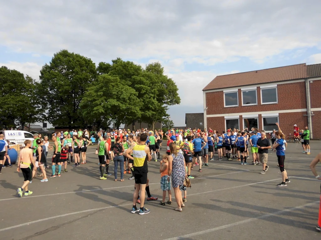 Teilnehmer eines Lauf-Events versammeln sich auf einem Parkplatz vor einem Gebäude, umgeben von Bäumen. Viele tragen Sportkleidung in verschiedenen Farben, während Zuschauer und Familienangehörige zuschauen.