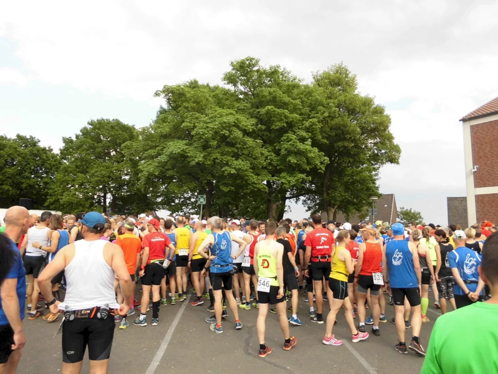 Teilnehmer eines Laufwettbewerbs versammeln sich auf einem Parkplatz, um sich auf den Start vorzubereiten. Die Menge besteht aus Läufern in bunten Sportoutfits, umgeben von Bäumen und Gebäuden.