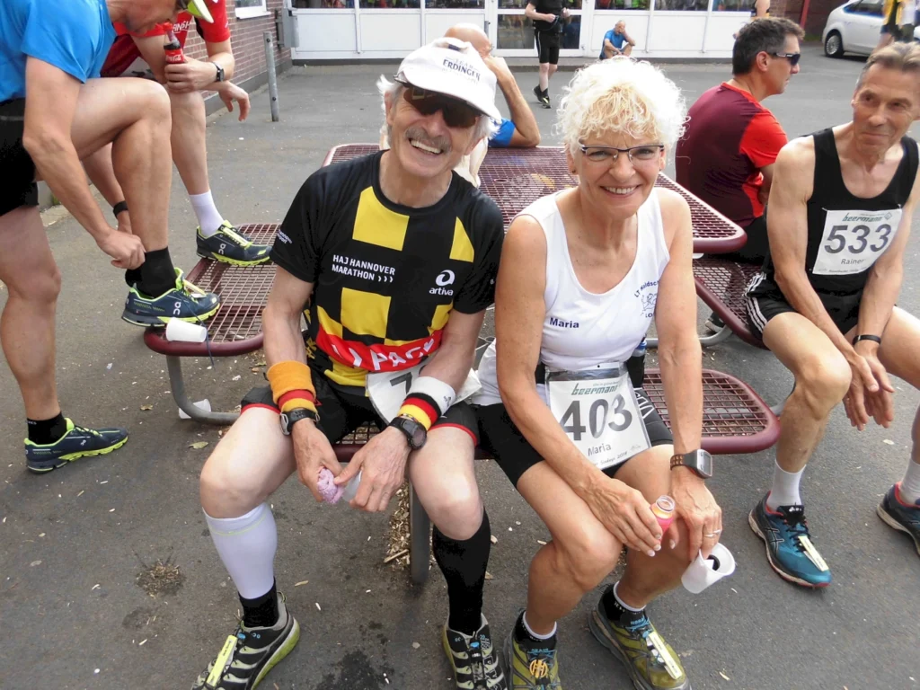 Teilnehmer des Marathons entspannen sich in der Pause. Ein älterer Mann in einem gelb-schwarzen Shirt und eine Frau mit lockigem Haar in einem weißen Shirt sitzen auf einer Bank, während andere Läufer im Hintergrund ihre Ausrüstung anpassen. Beide tragen Startnummern und scheinen sich auf den Wettkampf vorzubereiten.