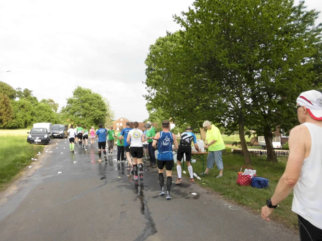 Läufer nehmen an einem Wettkampf teil, während sie an einer Verpflegungsstation vorbeikommen. Im Hintergrund sind Bäume und Fahrzeuge zu sehen, und die Straße ist mit Müllresten gesäumt.