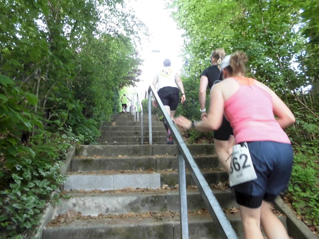 Läuferinnen steigen eine Treppe im Freien, umgeben von Bäumen und Pflanzen, während sie an einem Wettkampf teilnehmen.