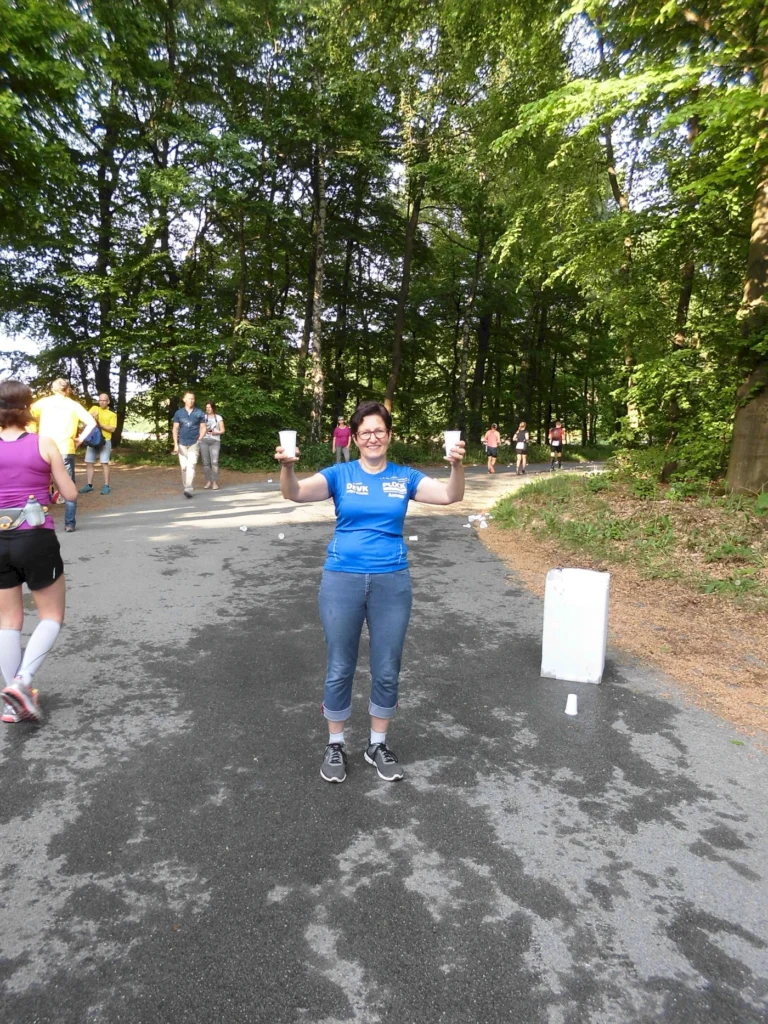 Frau in blauer Sportkleidung steht auf einer Laufstrecke im Wald und hält zwei Becher in der Hand, während Teilnehmer im Hintergrund vorbeilaufen. Ideal für Veranstaltungen oder Marathon-Highlights.